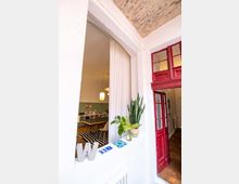 Ein Innenraum mit einem offenen Durchgang, der von einer roten Holztür und einer Fensteröffnung mit Vorhang eingefasst ist. Im Vordergrund befinden sich Pflanzen und Dekorationsobjekte, im Hintergrund ein Raum mit einem Holztisch, Stühlen, einer gestreiften Teppichmatte und in warmem Licht beleuchteten Pendelleuchten.