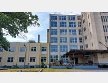 Industriegebäude mit hellbeiger und weißer Fassade, bestehend aus einem mehrstöckigen Hauptteil mit zahlreichen Fenstern und einem niedrigeren Seitentrakt. Im Vordergrund befinden sich ein überdachter Ladebereich, Paletten und weitere industrielle Materialien. Ein Baumrahmen und ein Schornstein sind sichtbar.