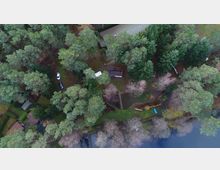 Luftaufnahme eines bewaldeten Grundstücks mit hohen Bäumen, einem kleinen Gebäude, einem Wohnwagen und geparkten Autos. Im unteren Bildbereich verläuft ein See, an dessen Ufer sich ein schmaler Pfad und ein blaues Kanu befinden.