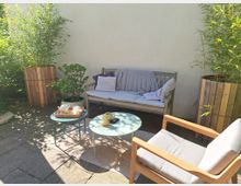 Kleiner, sonniger Innenhof mit Sitzmöbeln, bestehend aus einer Holzbank mit Polstern und einem Holzstuhl. Zwei runde Tische stehen in der Mitte, umgeben von hohen Bambuspflanzen in Holzkübeln und einem Pflasterboden.