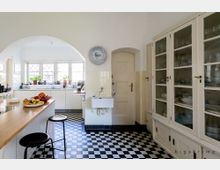 Helle, klassische Küche mit weißem Mobiliar und großen Glasvitrinen. Der Raum verfügt über ein Schachbrettmuster auf dem Fußboden, eine hölzerne Arbeitsfläche mit Hockern, ein Spülbecken neben einer Tür und große Fenster mit dekorativen Sprossen sowie Pflanzen auf der Fensterbank.