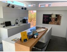 Moderner Küchenraum mit weißen Schränken und einer Marmor-Arbeitsplatte. Auf der Wand hängt ein Gemälde mit der Aufschrift "Where is Bud?", und auf der Holzbar im Vordergrund liegen Prospekte und Dekorationsobjekte.