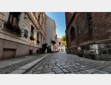Pflastersteingasse in einer Altstadt mit historischen Gebäuden; links eine Hausfassade mit Blumenkästen an den Fenstern, rechts eine Kirche aus roten Ziegelsteinen. Im Hintergrund ist ein weiteres Gebäude mit heller Fassade und ein Baum zu sehen.