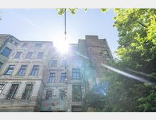 Fassade eines mehrstöckigen Altbaus mit hellen, leicht verwitterten Mauern und roten Fensterrahmen; im Vordergrund hängen Lichterketten, und die Szene wird von Sonnenschein und umgebenden Bäumen ergänzt. Unten ist ein Schild mit der Aufschrift „Clärchens Ballhaus“ sichtbar.