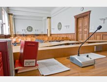 Ein Sitzungssaal mit Holzvertäfelung, über den ein Rednerpult mit Mikrofon, Gesetzesbüchern und Papieren im Vordergrund zu sehen ist. Im Hintergrund befinden sich hölzerne Sitzreihen, Säulen und eine große Holztür.