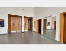 Ein einfacher Innenraum mit zwei großen Holztüren in der Mitte, flankiert von glatten Wänden in Weiß. Rechts hängt ein Spiegel, links befindet sich ein Gemälde an der Wand. Ein Desinfektionsspender und ein Feuermelder sind an der Wand montiert.