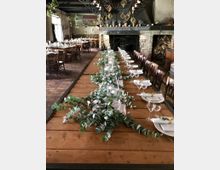 Langer, gedeckter Holztisch in einem rustikalen Raum mit Steinmauern und einem großen offenen Kamin. Der Tisch ist mit Eukalyptus-Zweigen, Kerzen und Gläsern dekoriert, während im Hintergrund weitere gedeckte Tische und ein Kaminholzstapel zu sehen sind.