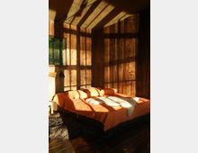 Schlafzimmer in einer Holzkabine mit warmem Tageslichteinfall. Ein Bett mit orangefarbener Bettwäsche und einem weißen Fellüberwurf steht vor holzvertäfelten Wänden, daneben ein Fenster, das Sonnenlicht ins Zimmer lässt.