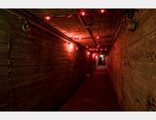 Langer, schmaler Betonflur mit rot leuchtenden Lampen an Decke und Wänden. Graffitis und Leitungen sind an den Wänden sichtbar, im Hintergrund führen Treppen zu einem beleuchteten Ausgang.