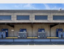 Industriegebäude mit mehreren Ladebuchtüren (43–46) und Metallschutzvorrichtungen, darüber Glasbausteinfenster entlang der oberen Fassade. Die Umgebung umfasst einen asphaltierten Bereich mit markierten Parkflächen.