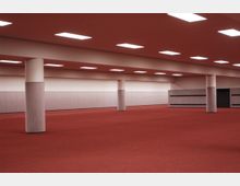 Ein großer, leerer Innenraum mit roten Teppichboden, weißen Wänden und mehreren quadratischen Deckenleuchten. Der Raum wird von mehreren runden Säulen und einem umlaufenden Streifenmuster an den Wänden strukturiert.