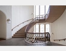 Großes Treppenhaus in einem historischen Gebäude mit geschwungener Treppe, verziertem Metallgeländer und großen Fenstern, die viel Tageslicht hereinlassen. Wände und Boden sind schlicht gestaltet, mit einem hohen weißen Treppenhausraum.