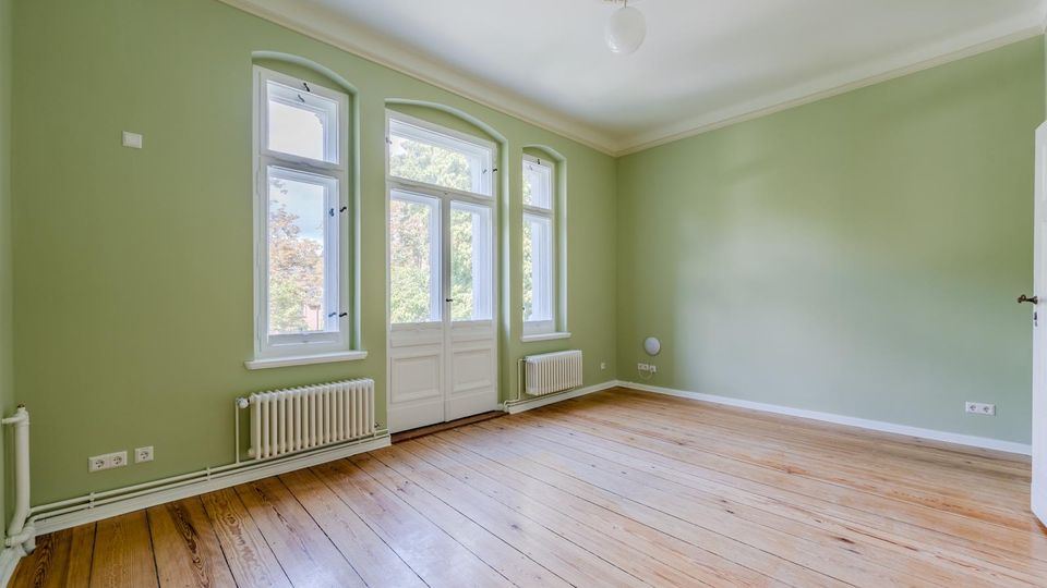 Heller, leerer Wohnraum mit hellgrünen Wänden, hohen Decken mit Stuckrosette und breiten Holzdielen. Drei hohe Fenster und eine verglaste Flügeltür lassen Tageslicht herein; unter den Fenstern stehen weiße Heizkörper.