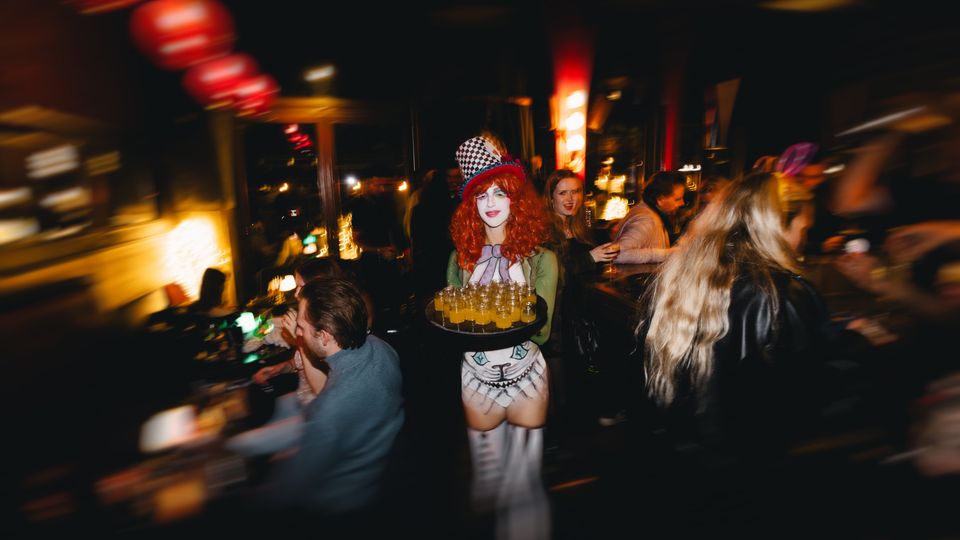 Innenaufnahme einer dunklen, belebten Bar oder eines Nachtclubs mit warmen Lichtquellen, roten Lampions an der Decke und einer langen Theke. Der Raum wirkt eng und ist von zahlreichen Gästen umgeben, mit glänzenden Oberflächen und reflektierenden Fenstern im Hintergrund.