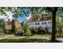 Straßenszene mit zwei zweistöckigen Wohnhäusern im Vordergrund, eines in weiß mit roten Fensterläden, das andere in gelb. Die Umgebung ist von Bäumen, Gärten und parkenden Autos auf einer kleinen Straße geprägt. Ein Straßenschild mit der Aufschrift „Juttastraße“ ist sichtbar.