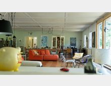 Weitläufiges Wohnzimmer mit heller Atmosphäre, einem großen roten Sofa im Vordergrund, einer langen Holztafel mit Stühlen im Hintergrund und großen Fenstern auf der rechten Seite. An den Wänden befinden sich Kunstwerke und Bücherregale, und die Decke ist mit sichtbaren Kabeln für Lampen gestaltet.