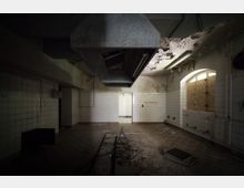 Ein verlassener Raum mit weiß gefliesten Wänden, heruntergekommenem Zustand und abblätterndem Putz. Deckenlüftung und Rohre sind sichtbar, das Fenster ist mit Holzplatten verbarrikadiert, und der Boden ist stellenweise beschädigt.