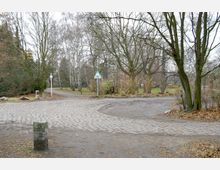 Kreuzung eines gepflasterten und eines unbefestigten Weges in einem Park, umgeben von kahlen Bäumen und Sträuchern. Im Hintergrund sind vereinzelte Grünflächen sowie ein Verkehrsschild zu sehen.
