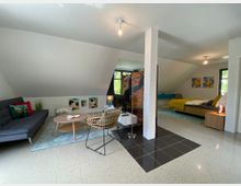 Großer, offen gestalteter Wohnraum in Dachgeschosslage mit einem modernen Sofa, einem niedrigen Holztisch und einem separaten Schlafbereich mit einem gelb bezogenen Bett. Der Raum ist hell, hat schräge Wände, dekorative Gemälde und kombiniert Wohn- und Schlafbereich durch eine kleine Trennwand.