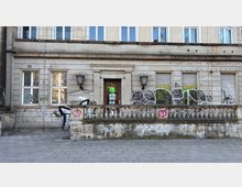 Straßenfassade eines mehrgeschossigen Steinbaus mit zentraler Eingangstür, zwei Wandlaternen und einer niedrigen Balustrade vor dem Gebäude. Mehrere Fenster mit Rollläden sowie großflächige Graffiti und Schmierereien sind an der Wand und am Sockel zu sehen.