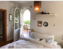 Schlafzimmer mit einem weißen Doppelbett und offener Balkontür, die zu einem gepflasterten Balkon mit Pflanzen und einem Metallstuhl führt. Die Wände sind weiß, dekoriert mit einem Regal, Bildern und einem auffälligen, bunten Kronleuchter.