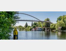 Eine geschwungene Fußgängerbrücke überspannt einen ruhigen Fluss, umgeben von Bäumen und Grünflächen. Am Ufer stehen kleine Häuser und Hausboote, während im Vordergrund ein gelb-schwarzer Pfeiler im Wasser zu sehen ist.