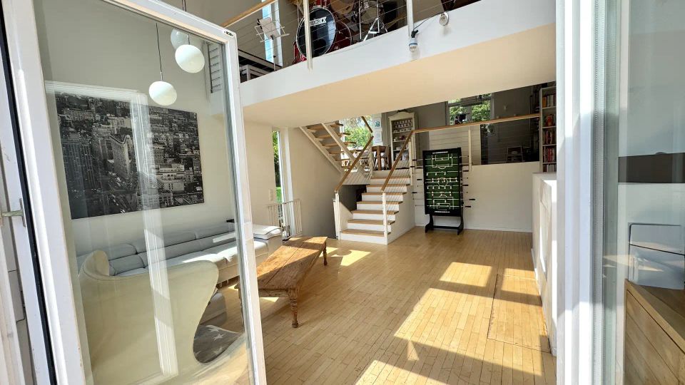 Heller, moderner Wohnraum mit hohen Decken, hellem Holzboden und großen Glasschiebetüren, die viel Tageslicht hereinlassen. Eine offene Treppe führt zu einer Galerie mit Geländer, auf der ein Schlagzeug steht; im Erdgeschoss sind Sitzbereich und ein Tischkicker zu sehen.