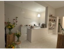 Heller, minimalistischer Ladeninnenraum mit weißem Tresen und Thekenbereich, Pendelleuchten und einer Kaffee- bzw. Kassenstation im Hintergrund. An den Wänden und auf Holzpodesten stehen Blumenarrangements, rechts hängen Regalbretter mit kleinen Produkten und Tüten.