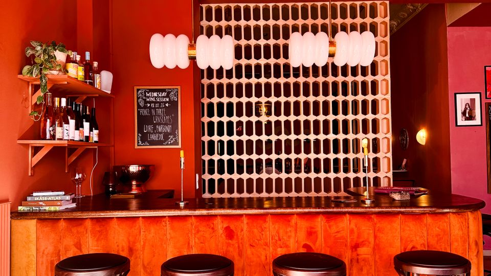 Interior of a small bar with vivid orange-red walls, a curved wooden counter, and four tall black bar stools on a tiled floor. Two pendant lights hang above the bar, with wall shelves of bottles and a chalkboard menu beside a white perforated partition behind the counter.