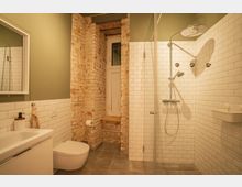 Badezimmer mit einer gemauerten Ziegelwand, einer weißen Fliesenumrandung und einer Dusche mit verglaster Kabine. Das Design kombiniert rustikale und moderne Elemente, mit einem Waschbecken, einer Toilette und einem kleinen Fenster mit weißem Rahmen.