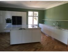 Moderne Küche mit weißer Einbauküche und grün gestrichenen Wänden. Ein zentraler Küchenblock mit Marmor-Arbeitsfläche, ein schwarzer Kühlschrank, ein eingebauter Backofen und ein großes Fenster sorgen für ein lichtdurchflutetes Ambiente; der Boden ist mit Fischgrätparkett ausgelegt.