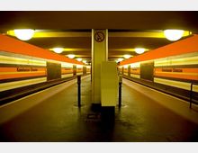 Ein U-Bahnhof mit zwei gegenüberliegenden Bahnsteigen, orange-gelb gestreiften Wänden und der Beschriftung „Konstanzer Straße“. Die Decke hat runde Deckenleuchten, und eine zentrale Säule mit einem Schild verbietet Rauchen.