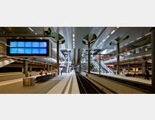 Großer, moderner Bahnhof mit mehreren Ebenen, hell beleuchteten Bahnsteigen und Rolltreppen, die zu höher gelegenen Plattformen führen. Ein großer Anzeigebildschirm zeigt die Gleisnummer, und im Hintergrund sind Geschäfte und Passagiere sichtbar.