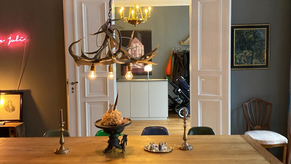 Esszimmer mit großem Holztisch und bunten Stühlen auf Fischgrätparkett, darüber eine auffällige Geweih-Hängelampe mit freiliegenden Glühbirnen. Im Hintergrund führen geöffnete weiße Flügeltüren in einen angrenzenden Raum mit Kronleuchter und Einbaumöbeln; an der Wand hängt ein gerahmtes Bild.