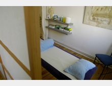 Behandlungsraum mit einer Liege, die mit einem weißen Tuch bedeckt ist und ein blaues Kopfkissen hat. An der Wand befinden sich zwei graue Regale mit medizinischen Utensilien, daneben ein abstraktes Wandbild; Holzboden und ein blauer Stuhl sind ebenfalls sichtbar.