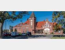 Backsteingebäude mit rotem Satteldach und einem zentralen Turm; typisch für historische Bahnhofsarchitektur. Vor dem Gebäude stehen parkende Autos, und im Vordergrund befinden sich ein Baum sowie eine Straße.