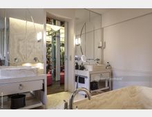 Modernes Badezimmer mit hellen Steinwaschbecken auf weißen Waschtischen, großen ovalen Spiegeln und verchromten Armaturen. Im Hintergrund ist ein begehbarer Kleiderschrank mit Spiegelflächen und rotem Sitzbereich sichtbar.