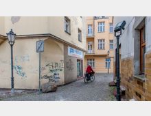 Enger gepflasterter Innenhof mit gelben Wohngebäuden, teils verputzten und teils mit Graffiti bedeckten Wänden. Eine Person auf einem Fahrrad fährt durch den Hof, neben einem Laternenmast und einem Verkehrsschild.