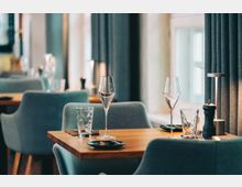 Modernes Restaurantinterieur mit Holztischen und gepolsterten, blaugrauen Stühlen; auf dem vorderen Tisch stehen Wein- und Wassergläser, Besteck, Teller sowie eine Pfeffermühle. Große Fenster mit dunklen Vorhängen lassen Tageslicht in den Raum.
