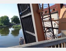 Geöffnete Klappbrücke aus Stahl, im Vordergrund ein Geländer aus Holz. Im Hintergrund ein ruhiger Fluss mit Bäumen und einem kleinen Gebäude mit rotem Dach.