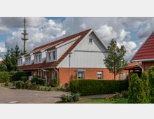 Zweistöckiges Wohnhaus mit roten Klinkerwänden im Erdgeschoss und weißer Holzverschalung im Obergeschoss, rotem Satteldach und gepflegtem Garten mit Sträuchern und Bäumen. Im Hintergrund ein hoher Funkmast und bewölkter Himmel.