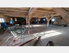 Großzügiger, hölzerner Innenraum eines historischen landwirtschaftlichen Gebäudes mit sichtbaren Holzbalken und Bretterboden. Mehrere alte Geräte und Maschinen, wie Pflüge und Körnermaschinen, sind ausgestellt; Fenster an den Backsteinwänden lassen Tageslicht herein.