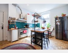 Helle, moderne Küche mit Parkettboden, einer Arbeitsinsel mit Barhockern und einer Wand aus türkisfarbenen Fliesen. Ausstattung umfasst einen Edelstahlherd, weiße Schränke, einen schwarzen Kühlschrank und ein großes Fenster, das viel Tageslicht hereinlässt; Zimmerpflanzen und hängende Küchenutensilien ergänzen die Einrichtung.