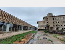 Ein verlassenes, teilweise verfallenes Industriegebäude mit Backsteinfassaden, großen Fensterfronten und rostigen Metalltüren. Der Boden besteht aus verwittertem Beton und Pflaster, teils überwuchert von Unkraut, unter wolkenbedecktem Himmel.