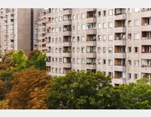 Wohnkomplex aus Beton mit mehreren Stockwerken und Balkonen, umgeben von dichtem, grünem und herbstlich gefärbtem Laub der Bäume. Die Fassaden wirken schlicht und funktional, typisch für städtischen Wohnbau.