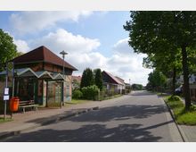 Straßenszene in einem Dorf mit einer Bushaltestelle im Vordergrund, die ein überdachtes Wartehäuschen mit Sitzbank und Abfallbehälter zeigt. Im Hintergrund sind niedrigere Häuser mit roten Dächern, Bäume und ein Bürgersteig zu sehen, die Straße ist ruhig und wenig befahren.