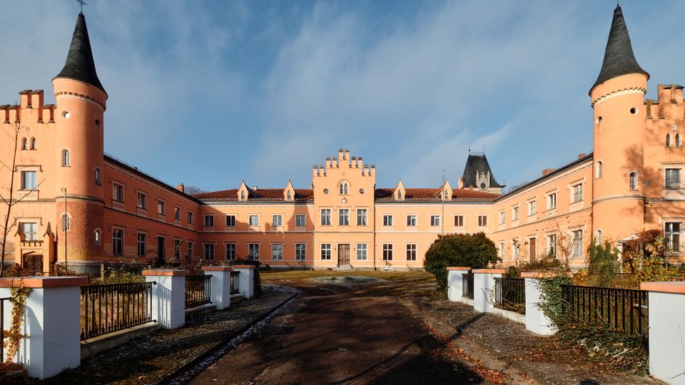 Innenhof eines schlossartigen, hellrosa Gebäudekomplexes mit zwei runden Ecktürmen und dunklen Spitzdächern sowie einer zentralen Fassade mit Zinnen. Ein breiter Weg führt durch den Hof, flankiert von niedrigen weißen Mauern mit Geländern, Sträuchern und kahler Vegetation unter blauem Himmel.