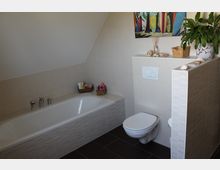 Badezimmer mit einer Badewanne links, einer wandmontierten Toilette rechts und hellen Fliesen an den Wänden. Dekorationselemente wie ein Bild, Muscheln und eine grüne Topfpflanze befinden sich auf einer Ablage.