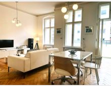 Wohnzimmer mit hellen Wänden und Holzboden im Fischgrätmuster. Im Raum stehen ein beiger Zwei-Sitzer, ein Fernseher auf einem weißen Lowboard, eine Esstischgruppe aus weißem Tisch und Stühlen, sowie hohe Fenster, die viel Tageslicht hereinlassen.