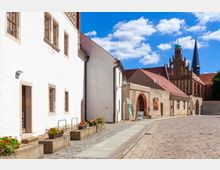 Historische Altstadtgasse mit Kopfsteinpflaster, flankiert von weißen und sandfarbenen Gebäuden mit Blumenbeeten. Im Hintergrund sind die roten Ziegeldächer und die Backsteinfassade einer mittelalterlichen Kirche mit Turmspitzen sichtbar.